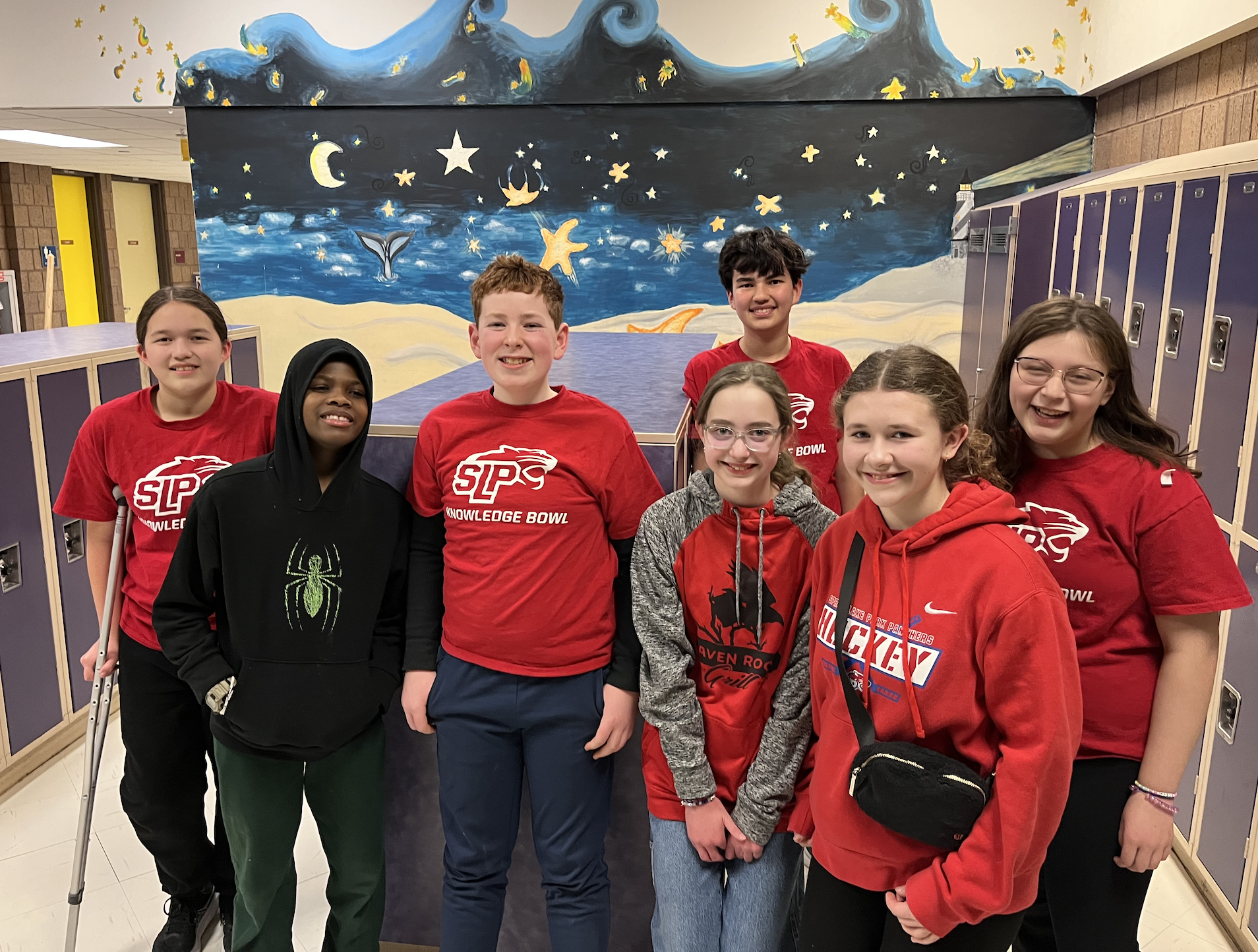 Group of smiling students wearing matching red t-shirts, posing in front of a painted mural with lockers nearby.